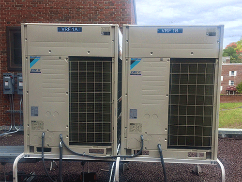 Two Daikin VRV IV units on rooftop beside brick building with visible piping connections.