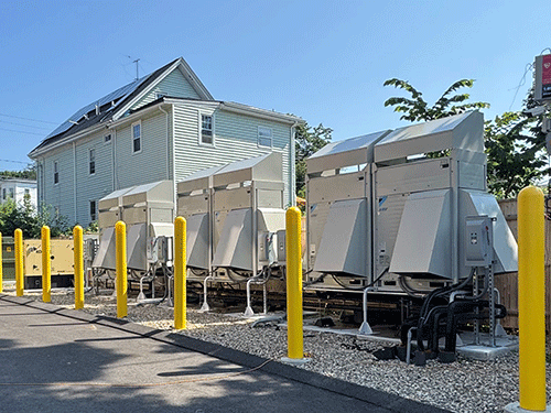 Outdoor view of several Daikin VRV heat pump condensing units installed on a gravel pad, lined with bright yellow protective bollards.
